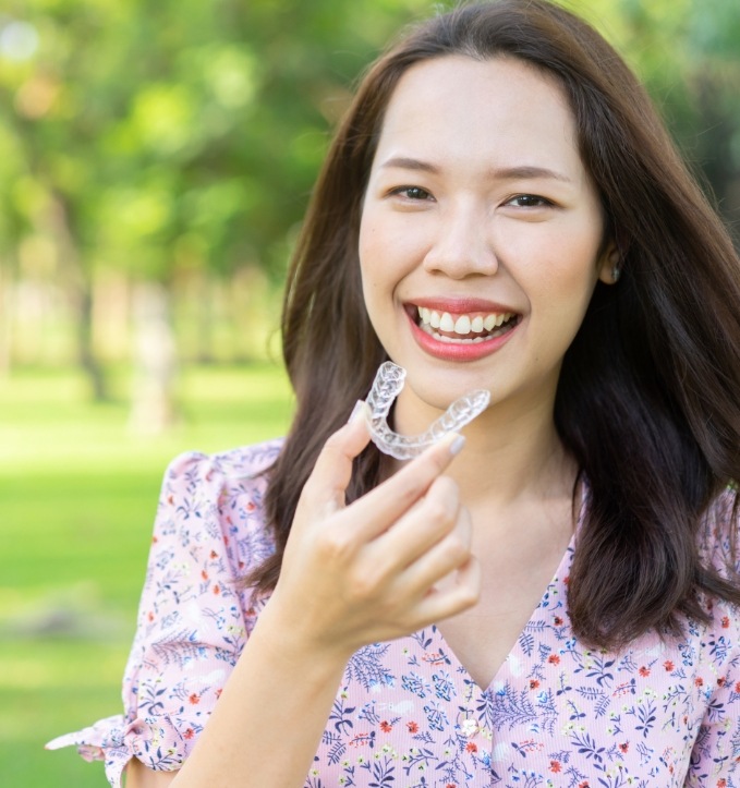 Woman smiling outdoors and holding an Invisalign clear aligner in Jonesboro